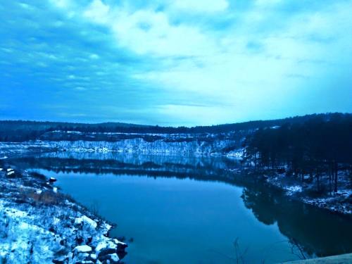 A serene winter landscape featuring a calm lake surrounded by snow-covered hills and a cloudy blue sky.