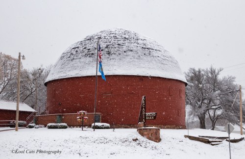 A round, red barn covered in snow, with an American flag and a sign reading "Arcadia Round Barn."