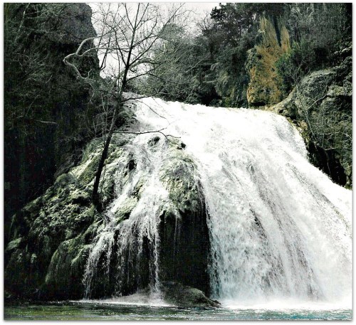 A cascading waterfall flows over rocky cliffs, surrounded by trees and lush greenery in a serene landscape.