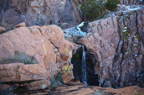 A small waterfall cascades over rocky terrain, surrounded by rugged cliffs and sparse vegetation.