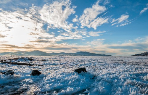 A frosty landscape with icy ground, scattered rocks, and a bright sky filled with clouds at sunrise.