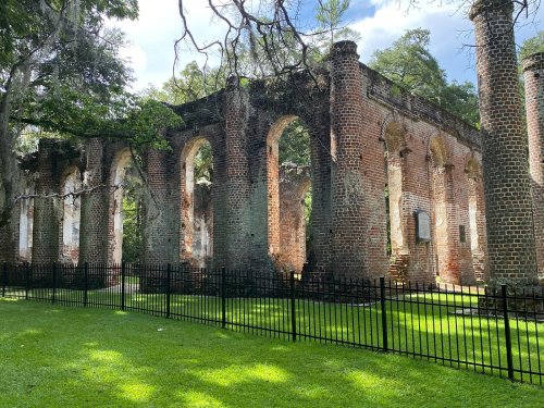 Ruins of a brick structure with arched windows, surrounded by green grass and a black fence under a blue sky.
