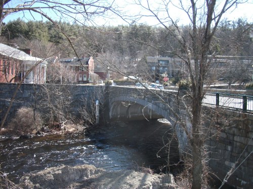A stone bridge spans a river, with trees and buildings in the background on a clear day.