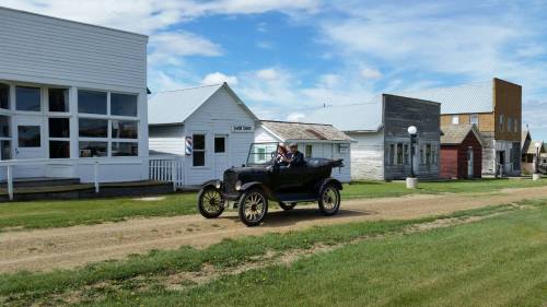A vintage car drives down a dirt road past historic buildings on a sunny day.