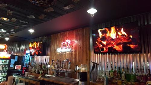 A cozy bar interior featuring wooden walls, illuminated screens showing a fire, and a variety of drinks on display.
