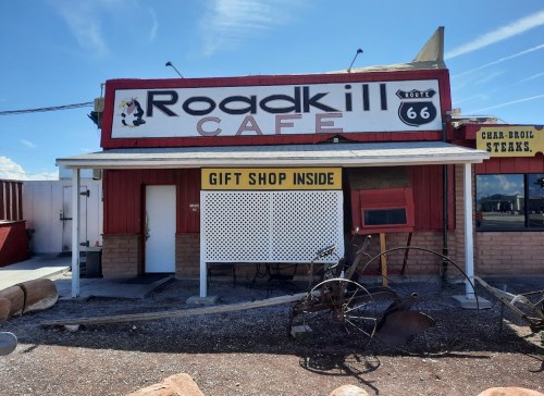 A roadside cafe with a sign reading "Roadkill Cafe" and "Gift Shop Inside," featuring rustic decor and Route 66 theme.