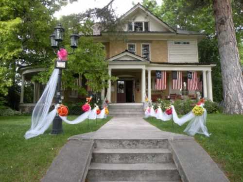 A charming house with a porch, decorated with flowers and draped fabric, flanked by American flags and greenery.