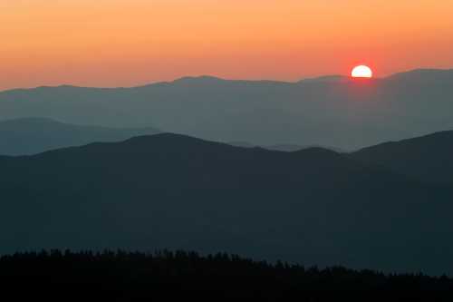 A serene sunset over layered mountains, casting warm orange hues across the sky and silhouettes of the peaks.