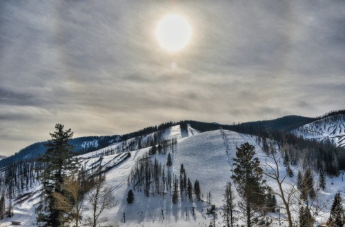A snowy landscape with rolling hills, tall trees, and a bright sun partially obscured by clouds in the sky.