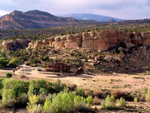 A scenic view of a rustic house nestled in a valley surrounded by rocky cliffs and lush greenery under a cloudy sky.