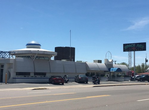 Exterior of the Space Age Restaurant, featuring a futuristic design and a sign, with cars parked outside.