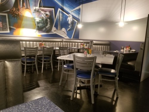 A modern diner interior with gray booths, blue accents, and space-themed wall art. Tables are set for dining.