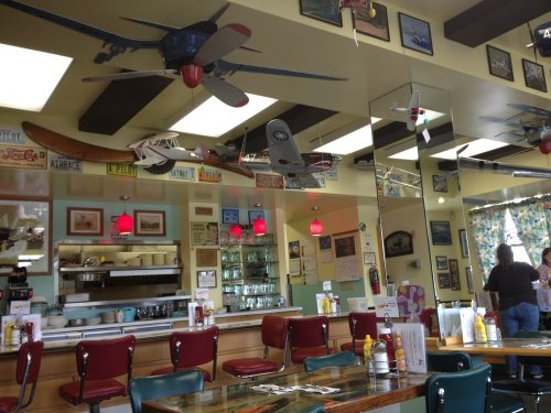 A retro diner interior with airplane decor, red pendant lights, and vintage signs, featuring tables and a counter.