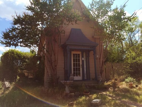 A quaint, small house with a blue roof, surrounded by greenery and a sunny sky.