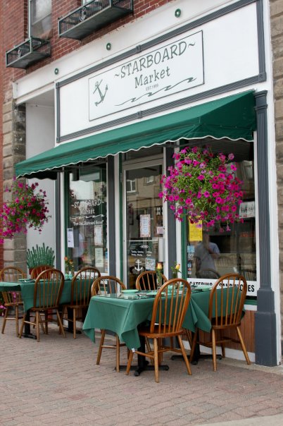Exterior of Starboard Market with green awnings, flower baskets, and outdoor seating on a brick sidewalk.