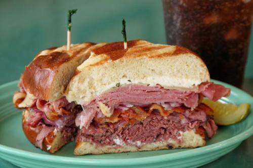 A hearty sandwich filled with layers of meats, topped with a toothpick, served with a drink on the side.