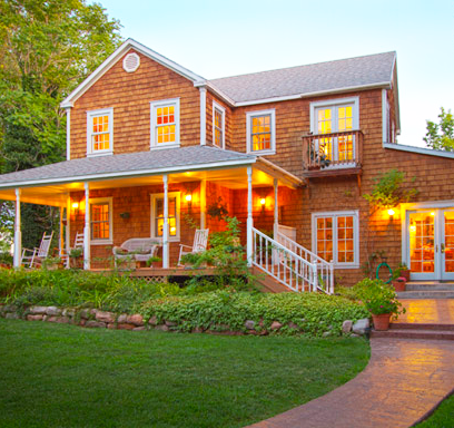 A charming two-story wooden house with a porch, surrounded by greenery and warm evening lighting.