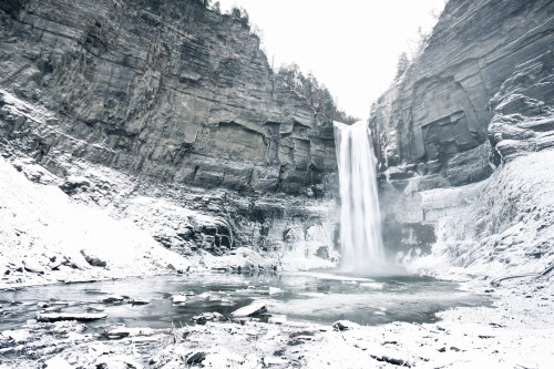 A snowy landscape featuring a tall waterfall cascading into a frozen pool, surrounded by rocky cliffs and trees.