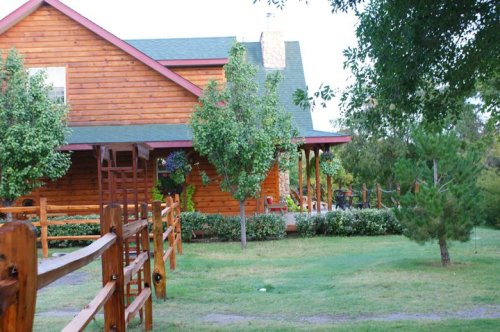 A rustic wooden house surrounded by trees and a green lawn, with a wooden fence in the foreground.