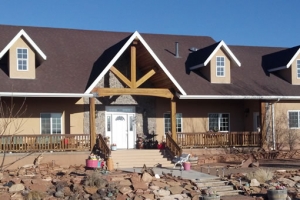 A large, two-story house with a brown roof, wooden porch, and stone accents, surrounded by a rocky landscape.