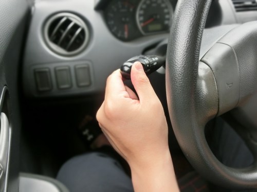 A hand gripping a car's turn signal lever, with the steering wheel and dashboard visible in the background.