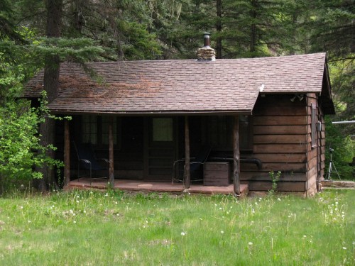 A rustic wooden cabin surrounded by trees, featuring a porch and a grassy area in front.