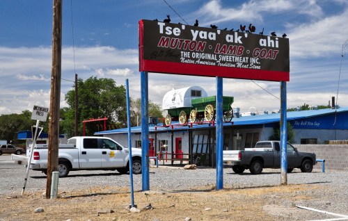 Sign for "Tse' yaa' ak' ahi" featuring mutton, lamb, and goat, with a blue building and parked trucks in the foreground.