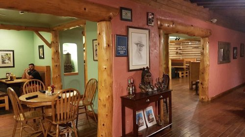 A rustic restaurant interior with wooden beams, tables, and framed artwork on colorful walls. A man sits at a table.