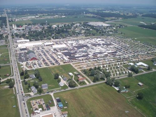 Aerial view of a large outdoor market surrounded by fields and a small town, with numerous parked cars and buildings.