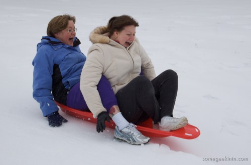 Two women sledding down a snowy hill, both with excited expressions, sitting on a red sled.