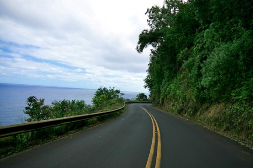 Winding road along a lush green hillside with ocean views under a cloudy sky.