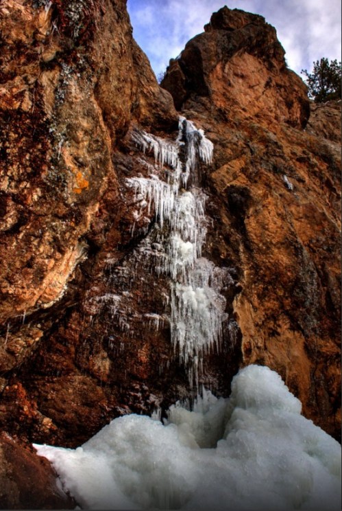 A frozen waterfall cascading down rocky cliffs, surrounded by ice and snow under a cloudy sky.