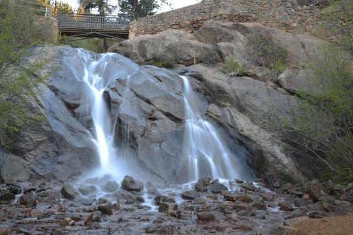 A serene waterfall cascades over rocky terrain, with a bridge visible in the background amidst lush greenery.