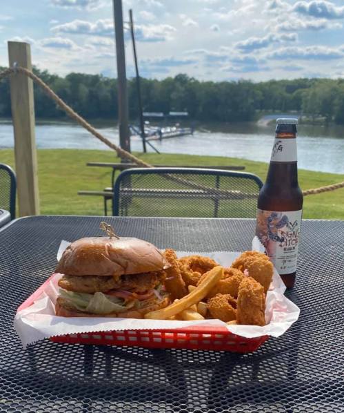 A burger with fries and chicken tenders, alongside a beer, set on a table with a river view in the background.