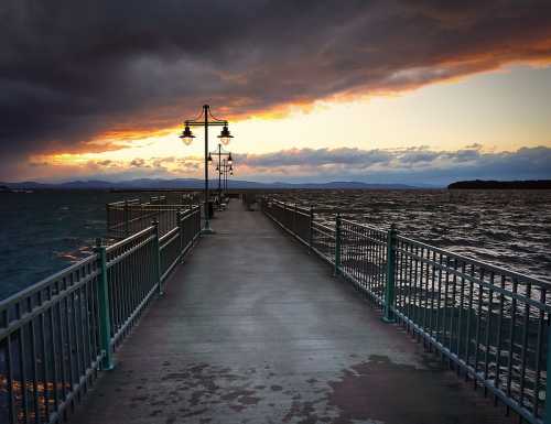 A wooden pier extends into a turbulent sea under a dramatic sunset with dark clouds and distant mountains.