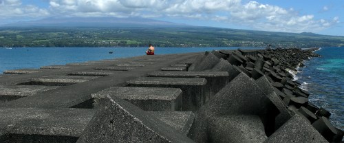 A person sits on a rocky pier extending into a blue ocean, with mountains and clouds in the background.