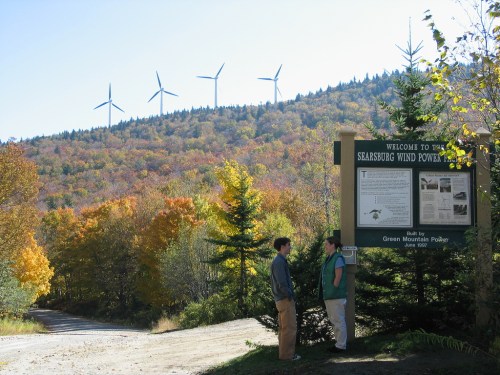 Two people stand near a sign for Searsburg Wind Power, with colorful autumn trees and wind turbines in the background.