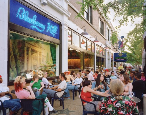 A lively outdoor scene at Blueberry Hill, with people socializing at tables under trees and neon lights.