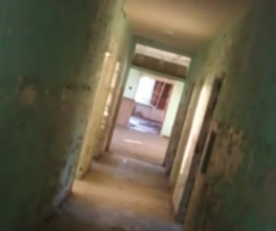 A dimly lit, abandoned hallway with peeling green walls and doorways leading to empty rooms.