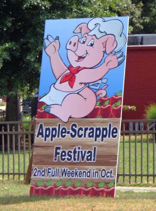 A colorful sign for the Apple-Scrapple Festival featuring a cheerful pig character, promoting the event in October.