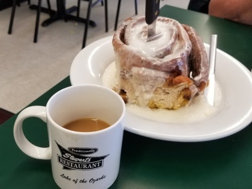 A large cinnamon roll with icing sits on a plate next to a cup of coffee at a restaurant.