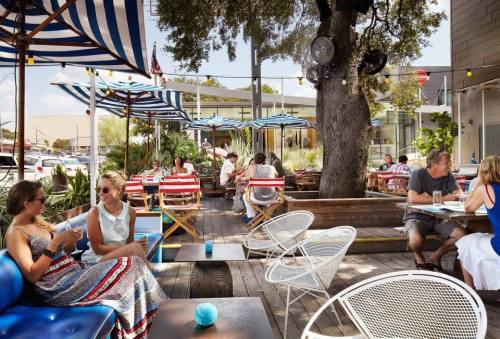 Outdoor dining area with striped umbrellas, wooden seating, and people enjoying drinks and conversation in a lively atmosphere.