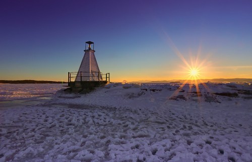 A lighthouse on a snowy landscape at sunset, with vibrant colors and a starburst effect in the sky.