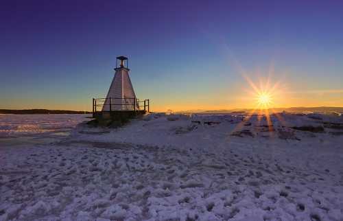 A lighthouse on a snowy landscape at sunset, with vibrant colors and a starburst effect in the sky.