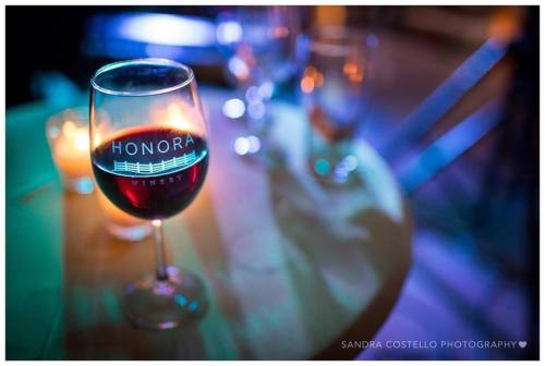 A glass of red wine sits on a table, illuminated by colorful lights, with a candle in the background.
