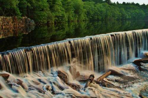 A serene waterfall cascades over rocks, surrounded by lush green trees and reflecting in calm water.