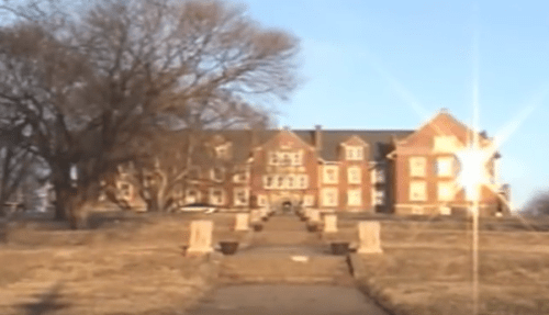 A large brick building with multiple windows, set against a clear sky, surrounded by bare trees and a pathway leading up to it.