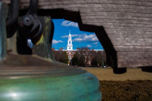 A bell in the foreground frames a view of a church steeple and blue sky with clouds in the background.