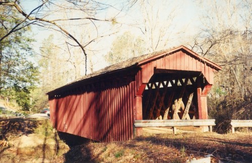 A red covered bridge stands over a small creek, surrounded by bare trees and a clear blue sky.