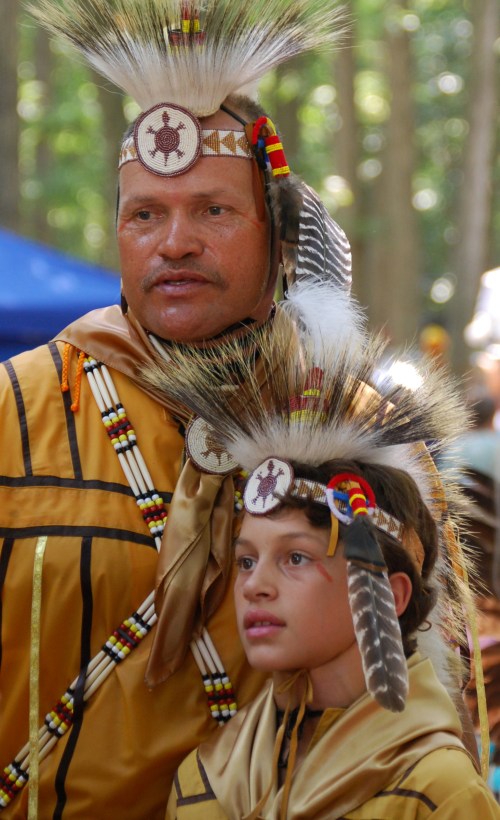 A man and a child in traditional Native American attire, adorned with feathered headdresses, standing in a forest setting.